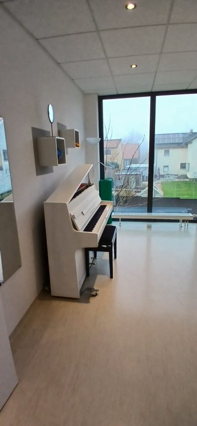 Raum 1: Ein Klavier steht in einem hellen Raum mit großen Fenstern und Blick ins Freie.