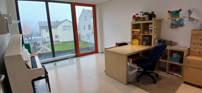 Raum 1: Heller Raum mit einem Schreibtisch, Regalen, einem Klavier und großen Fenstern.